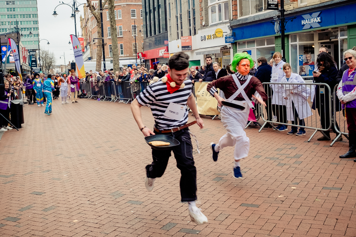 A crowd watching two men - dressed up as a Frenchman and an Oompa Loompa flipping pancakes and running on the high street